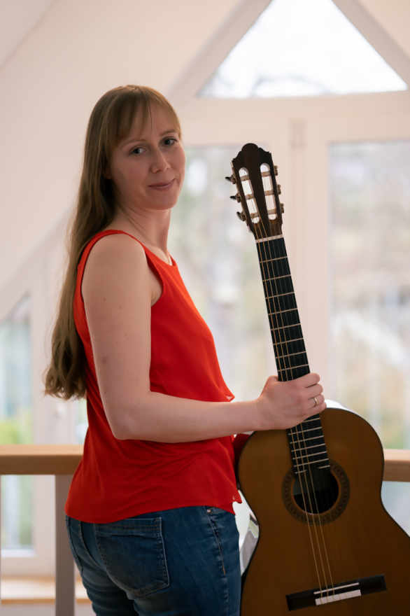 Claudia Kluck steht mit einer Gitarre innen vor mehreren Fenstern