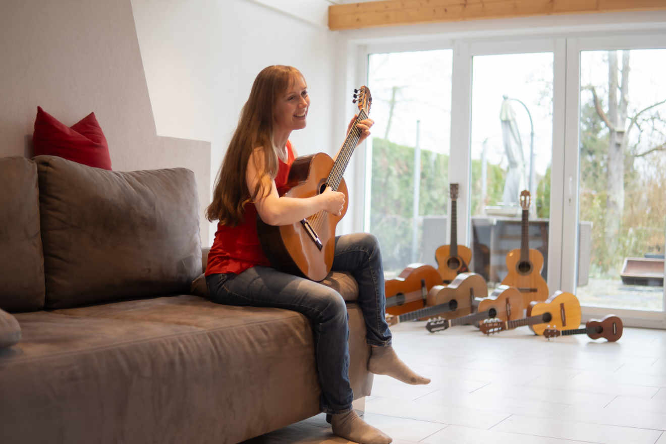 Claudia Kluck spielt Gitarre auf dem Sofa, im Hintergrund sind viele Gitarren zu sehen